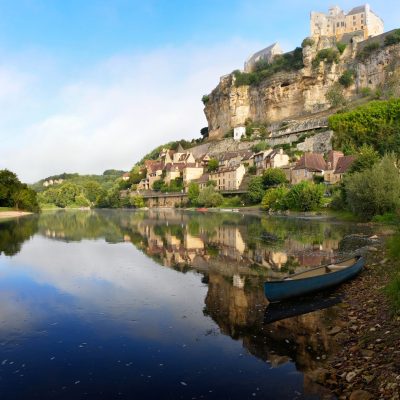 The beautiful village of Beynac-et-Cazenac with its characteristic Chateau de Beynac castle built upon the cliffs alongside Dordogne river in the early morning light with reflection in the water and a kayak in the foreground in the Perigord region in France.